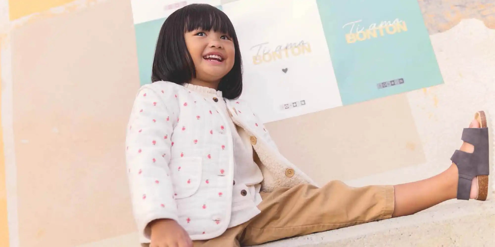 Un enfant portant une chemise blanche à pois et un pantalon beige est assis avec une expression joyeuse.